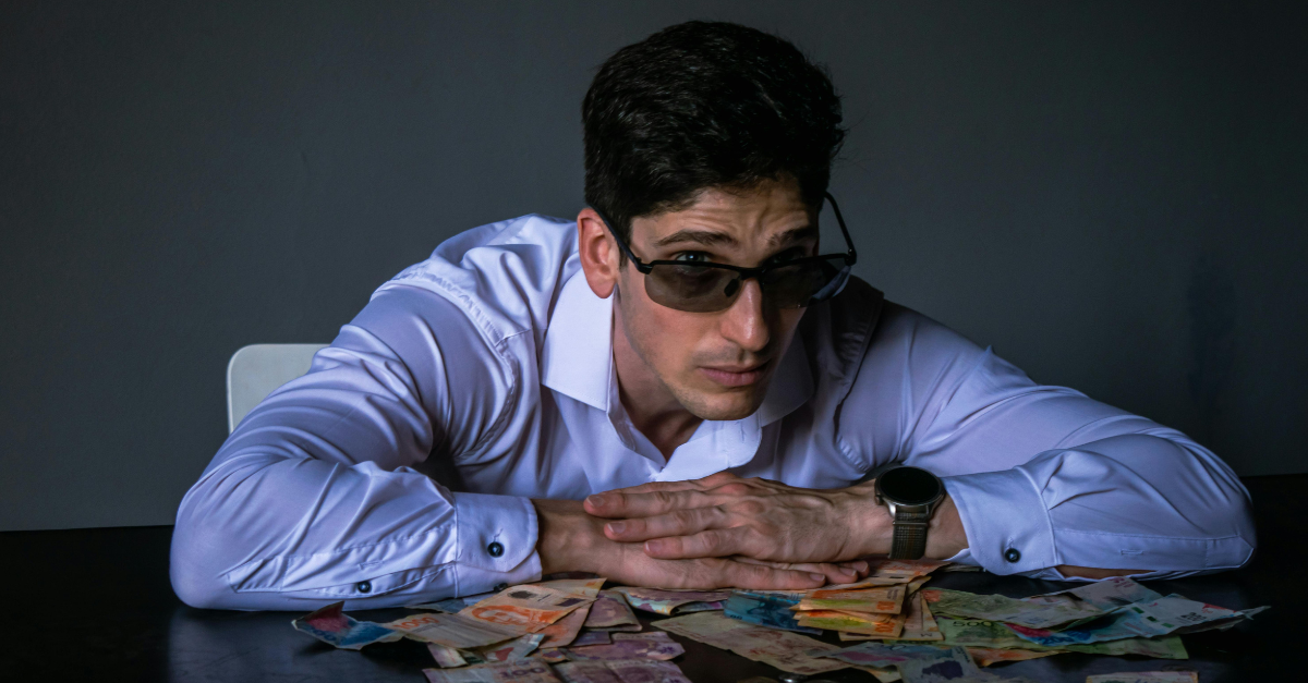 man-in-sunglasses-leaning-on-table-of-money