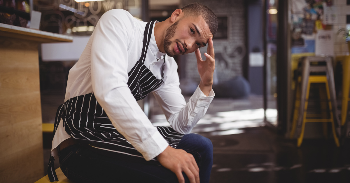 Upset young waiter sitting with headache while looking away at coffee shop