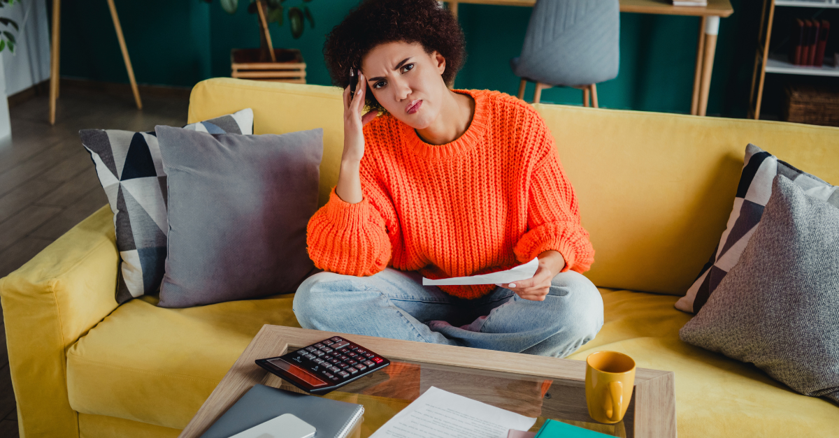A young woman sits on a yellow sofa at home, looking frustrated while reviewing bills