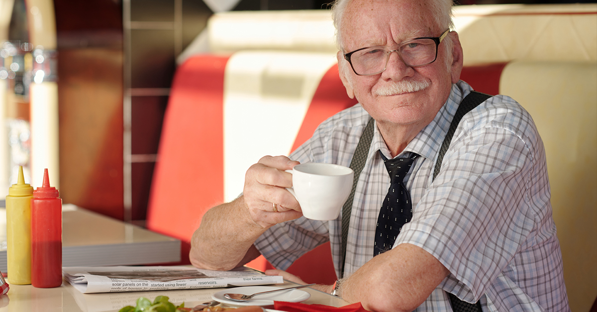 Portrait of elderly man with mustache sitting in diner, smiling while holding coffee cup, with food on table and red, white booth in background