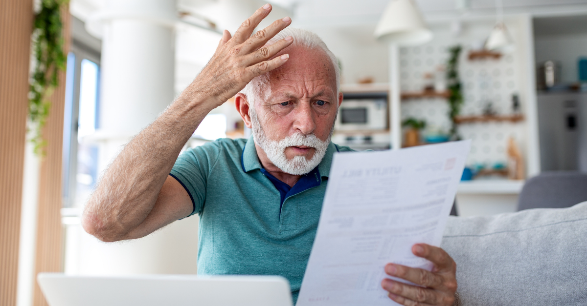 Worried senior man reading utility bill and using laptop at home.