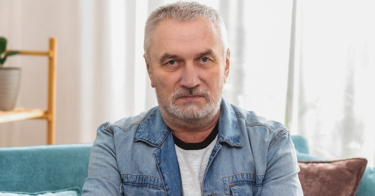 Angry senior man with gray hair and crossed arms looking at camera, sitting on sofa at home